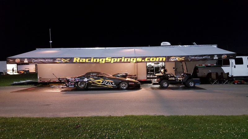 Race trailer awning lit up at night with a drag car parked underneath